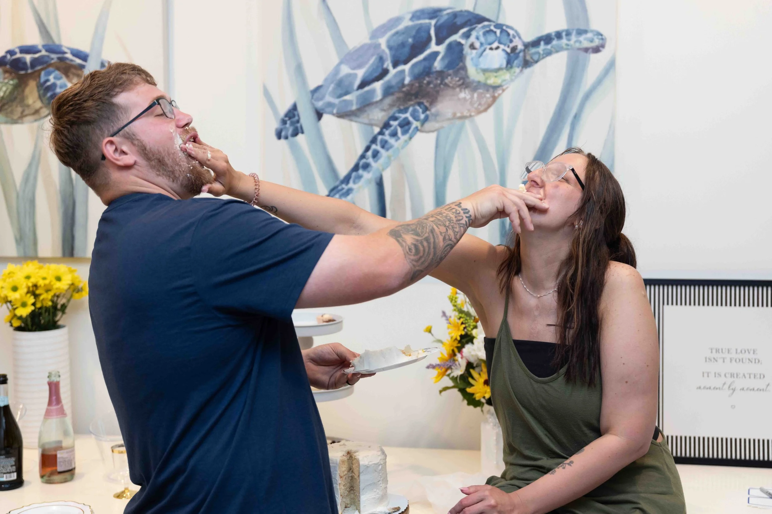 A man and woman sharing a cake at a celebration; the man appears to be playfully feeding cake to the woman, who is leaning forward with her mouth open. They are indoors surrounded by flowers and decorative jungle-themed wall art.