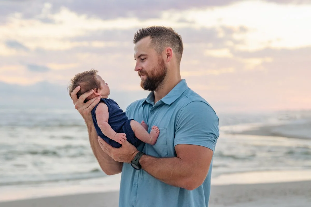 A man holding a newborn baby on the beach during sunset, gazing at the baby with affection.