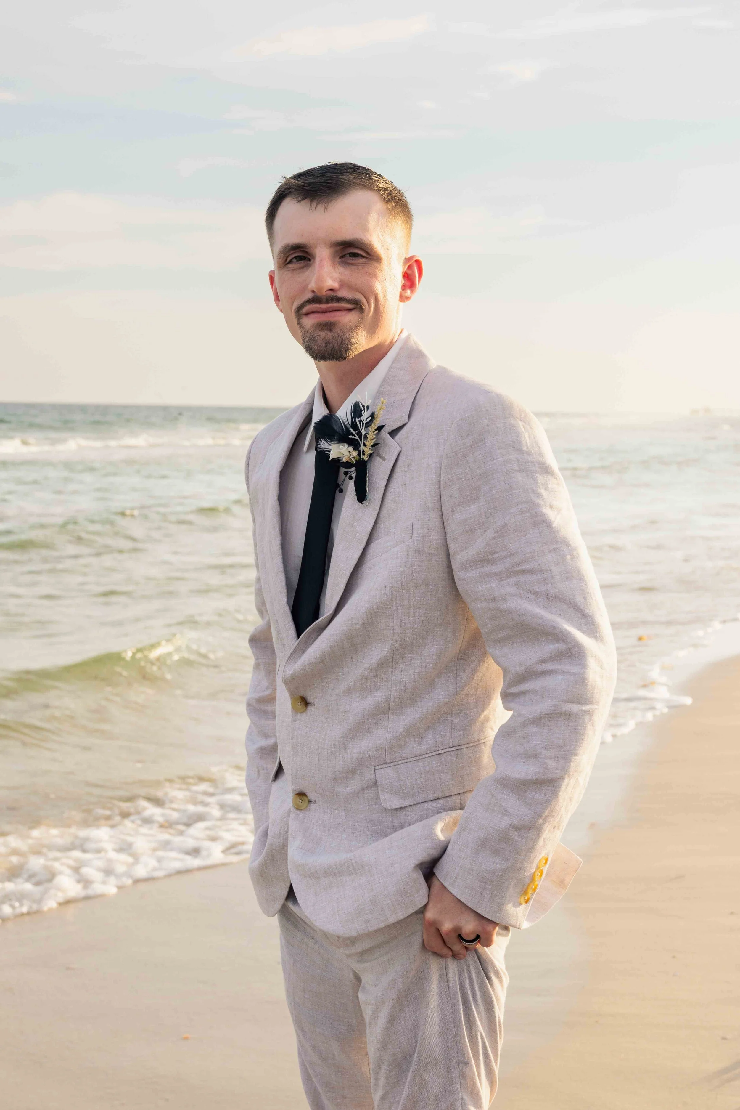 A man in a light-colored suit with a boutonniere, standing on the beach with the ocean in the background, during late afternoon or early evening.