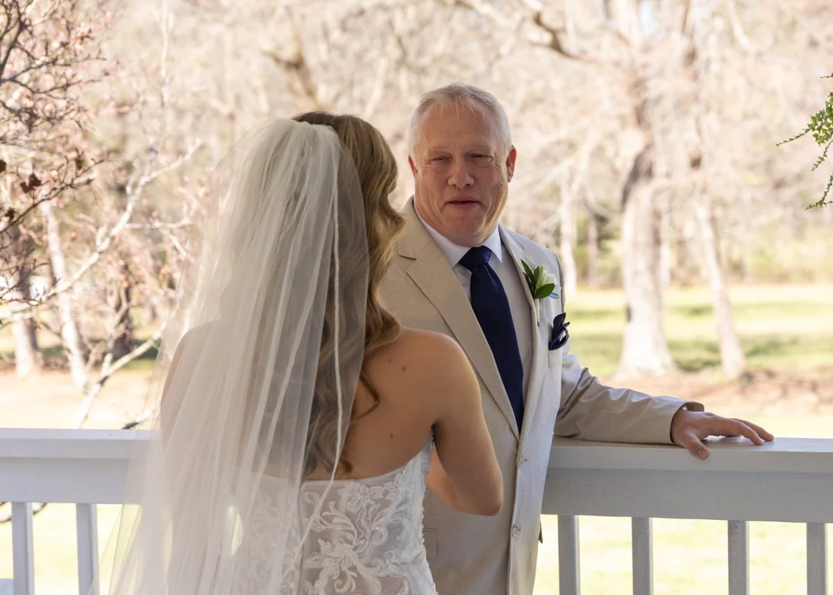 A bride and a man, likely her father, sharing a moment on a porch with trees in the background. The bride wears a white lace wedding dress and veil, and the man is dressed in a light-colored suit with a dark tie.