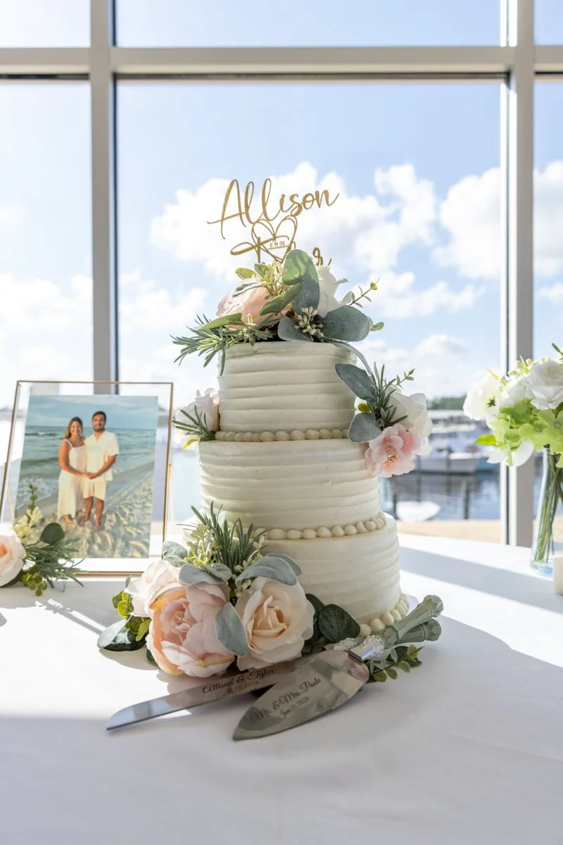 A three-tier white wedding cake decorated with pink and white flowers, greenery, and a gold "Allison" cake topper, surrounded by floral arrangements, a framed photo of a couple at the beach, and two cake serving knives, all set against a large window