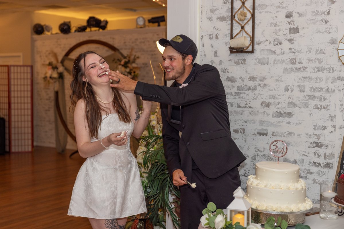A groom playfully feeding cake to a bride at a wedding reception with wedding cake and decorations in the background.