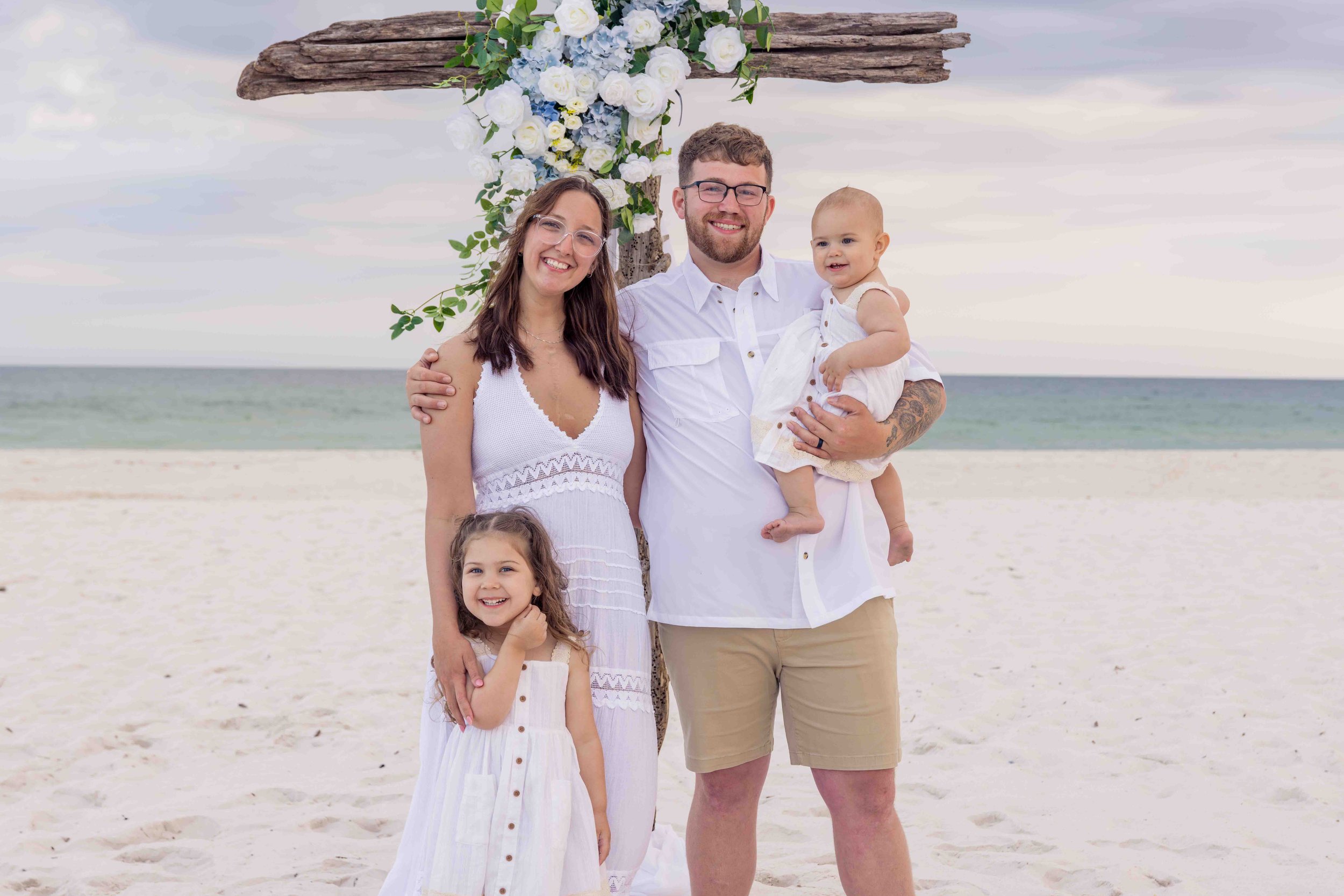 A smiling family of four standing barefoot on a beach in front of a decorative floral arch with white and blue flowers and green leaves, over a cloudy sky and calm ocean.