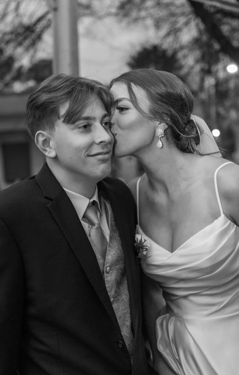 A black and white photo of a woman giving a kiss on the cheek to a man in a suit at a formal event, likely a wedding.