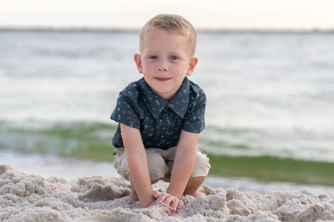 A young boy crouching on a sandy beach near the water, looking at the camera with a slight smile.