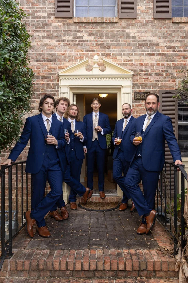 Six men dressed in matching blue suits and brown shoes, holding drinks, standing on the porch of a brick house.