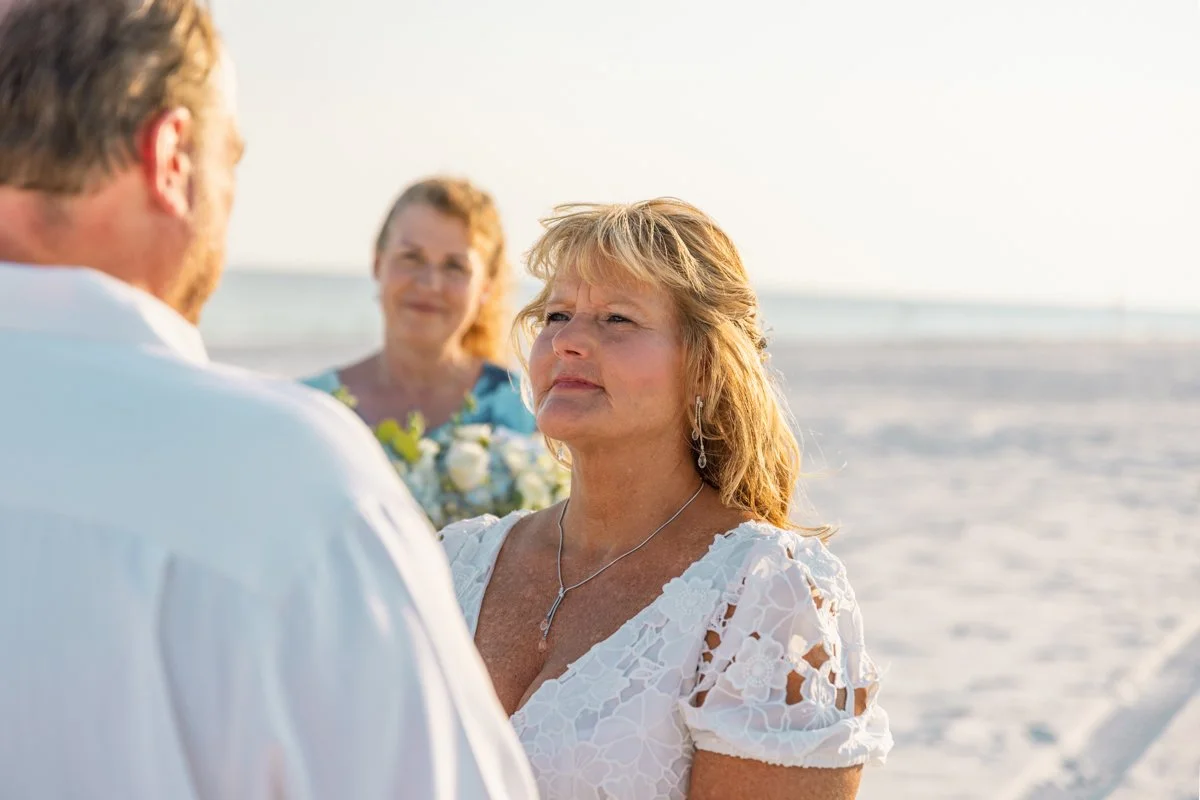 A woman at a beach wedding gazes at her partner during the ceremony, with an older woman holding a bouquet in the background.