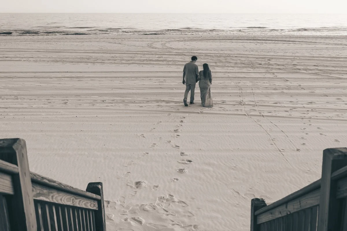 A man and woman walking hand in hand on a sandy beach, viewed from the top of wooden stairs.