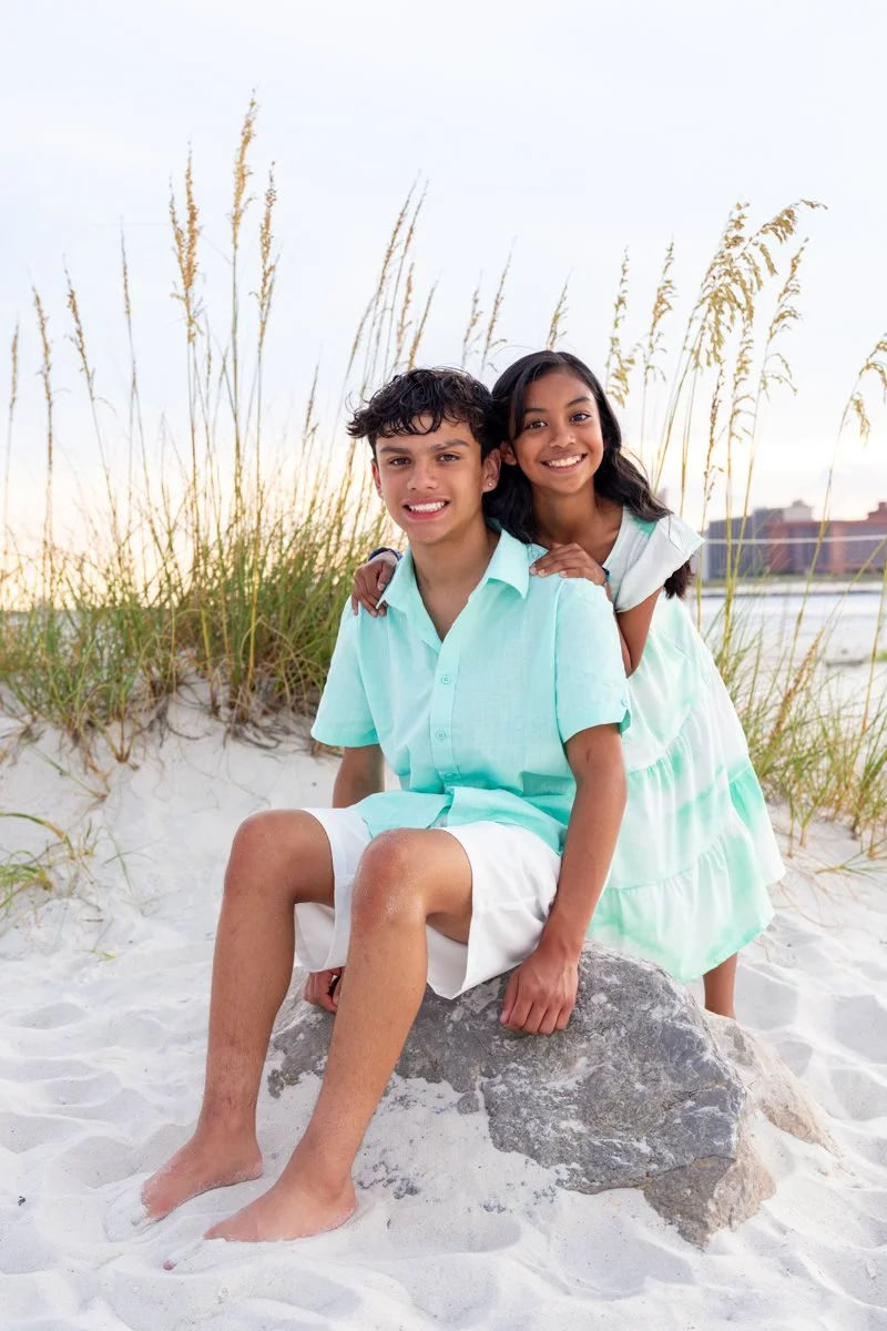 A boy and girl smiling on a beach, with the boy sitting on a rock and the girl standing behind him, both wearing light blue clothes, sandy dunes with beach grass and a body of water in the background.