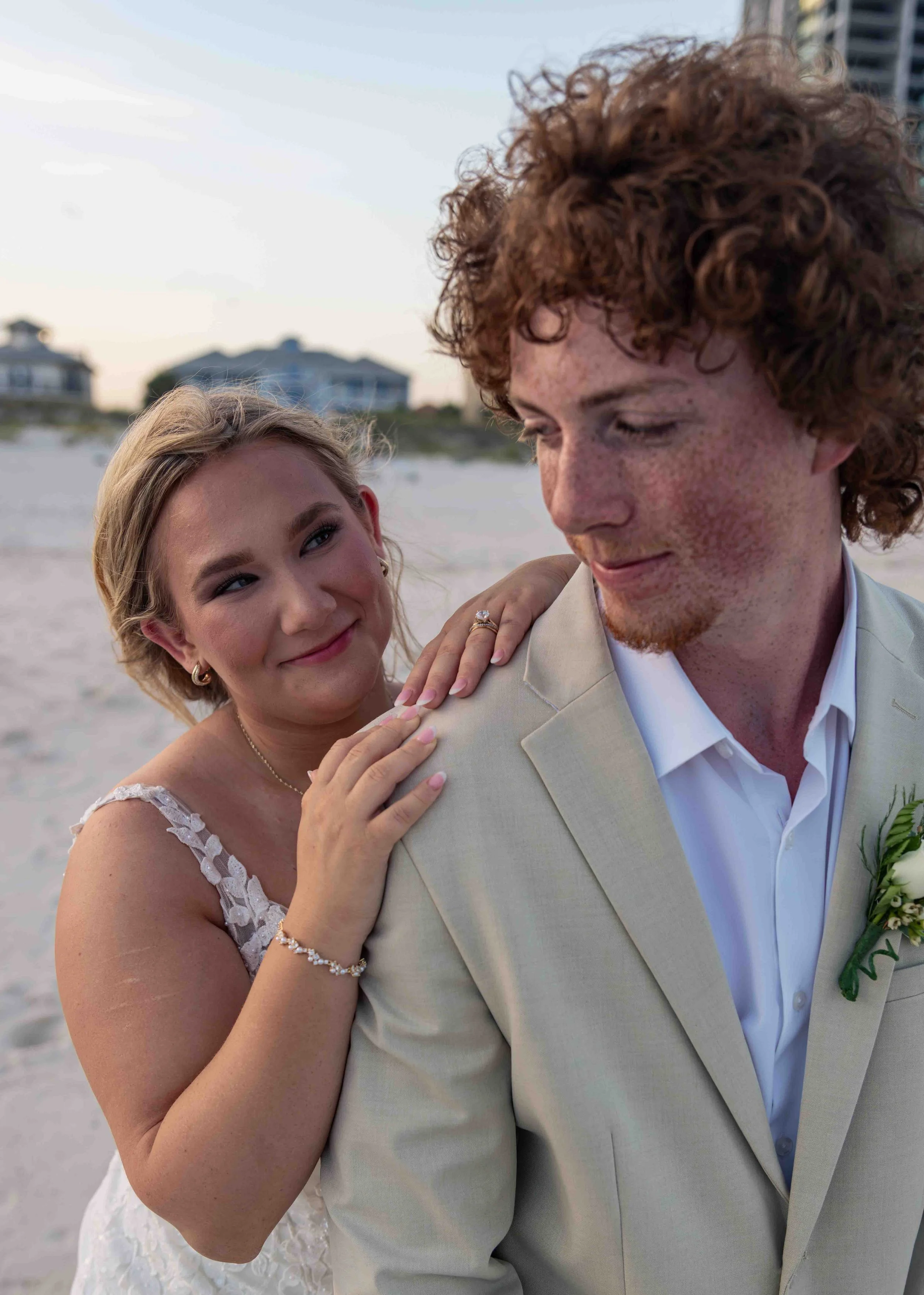 A bride and groom embrace on the beach at sunset, with the bride smiling and looking at the groom, who has curly red hair and freckles, wearing a light-colored suit. The bride has blonde hair and is wearing a white dress with lace details, and is hol