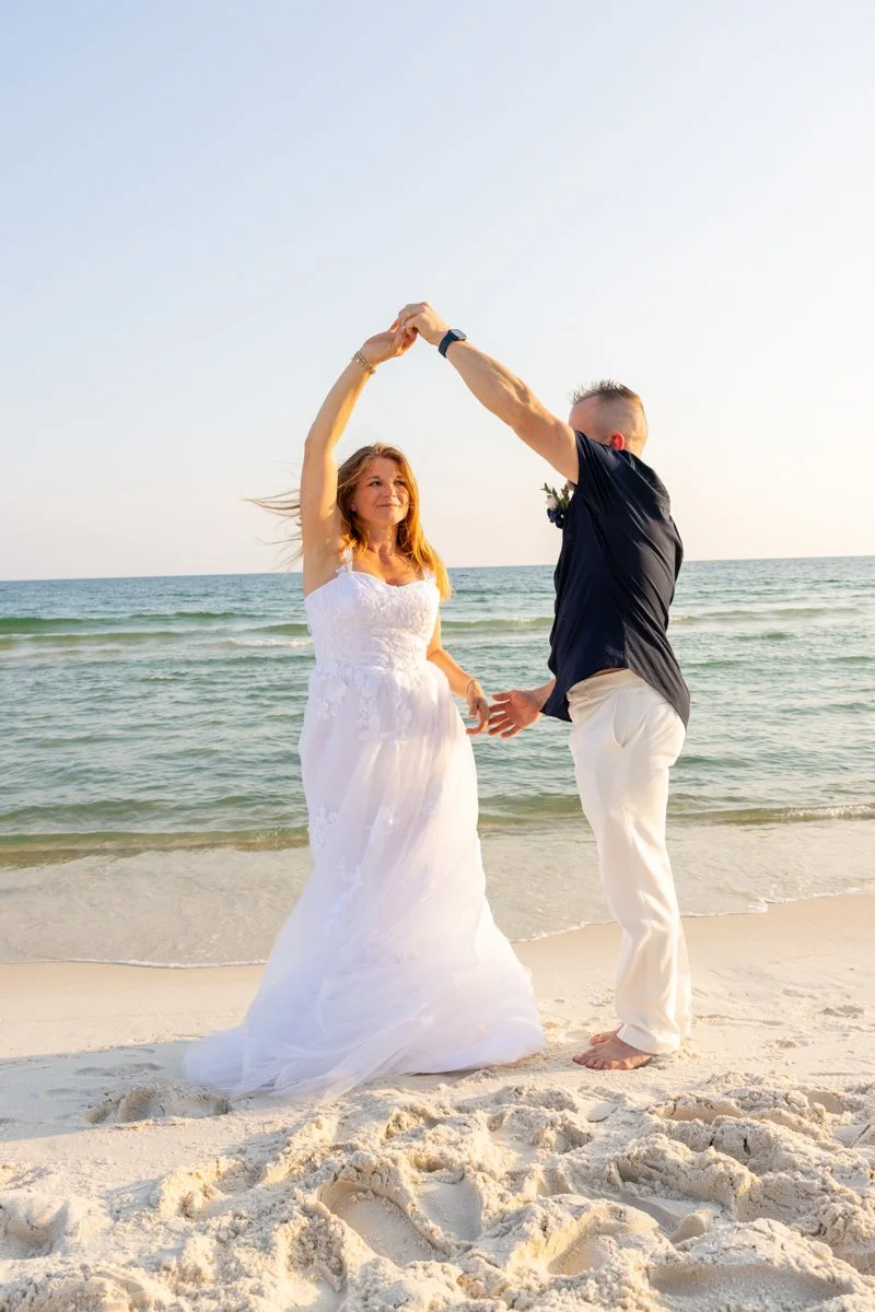 A couple dancing on the beach, the woman in a white wedding dress and the man in a black shirt and white pants, during sunset.