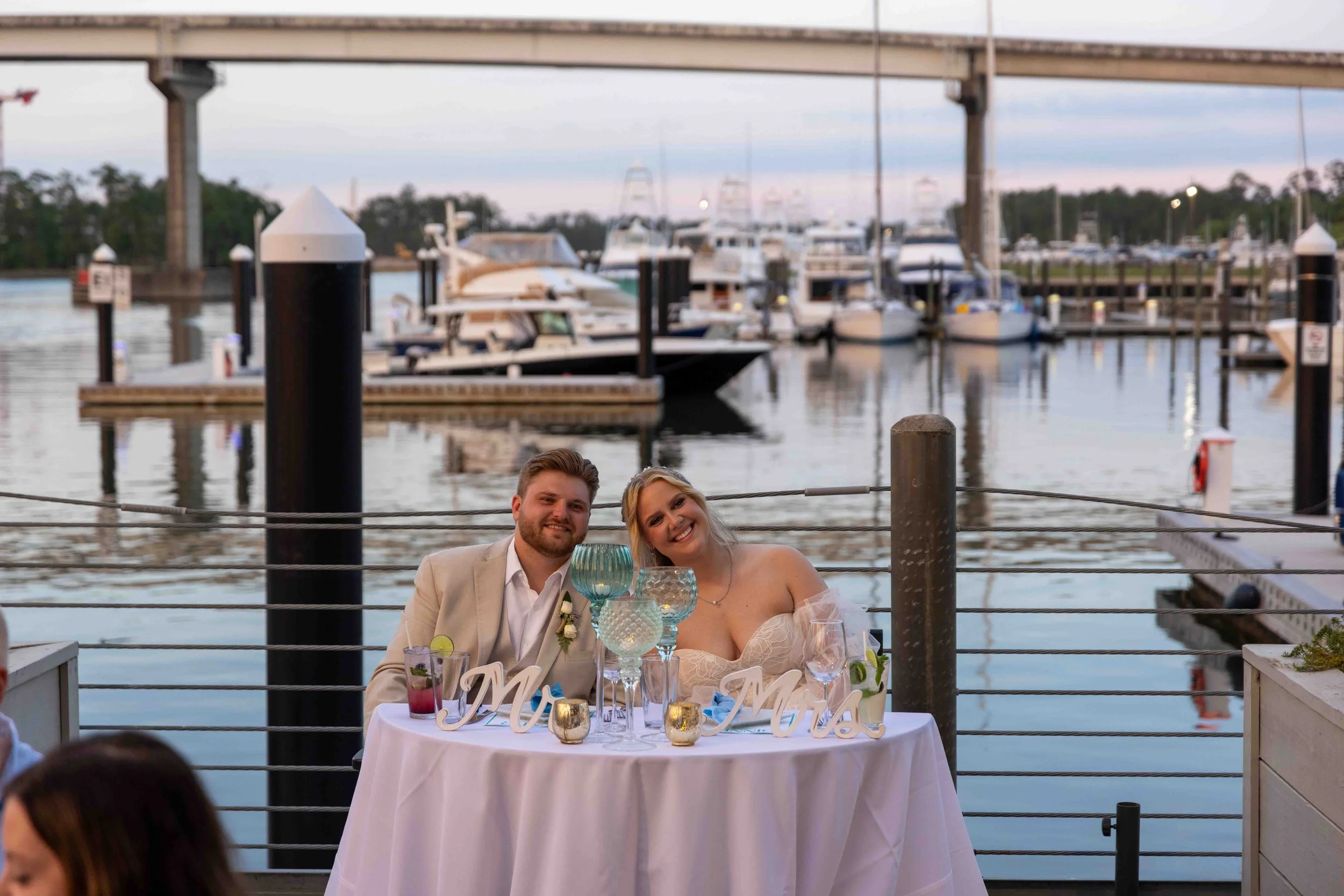 Wedding reception with a bride and groom sitting at a table by the water, enjoying their celebration with boats in the background.