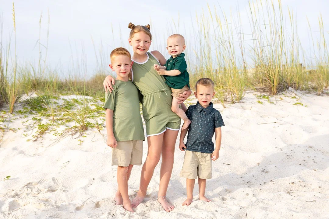 A girl holding a baby, with two boys, standing barefoot on a sandy beach with grass in the background, smiling
