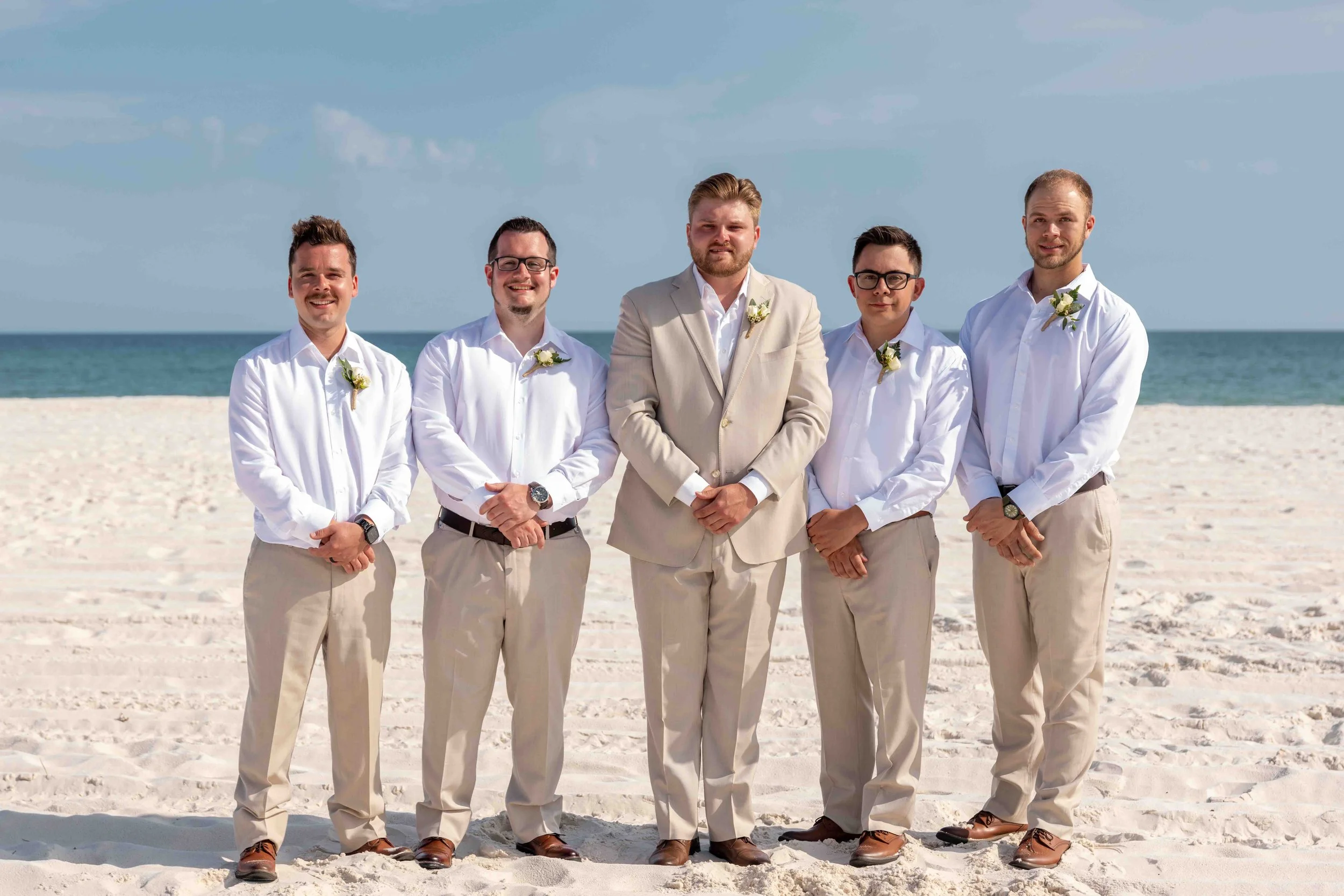 Group of five men dressed in light-colored suits and white shirts standing on the beach with the ocean and sky in the background.