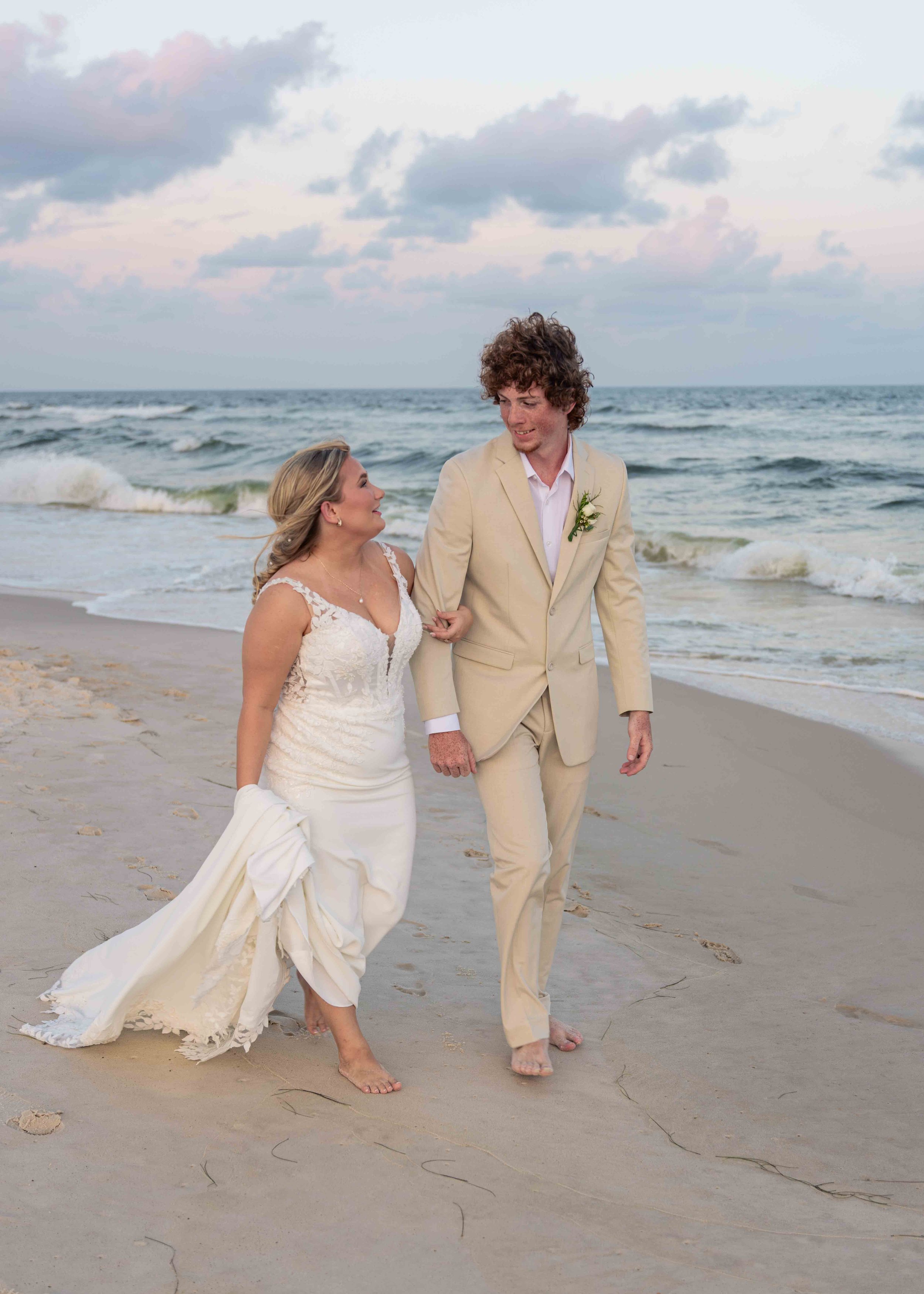 A bride and groom walking barefoot on the beach with ocean waves behind them, holding hands and looking at each other, dressed in wedding attire.