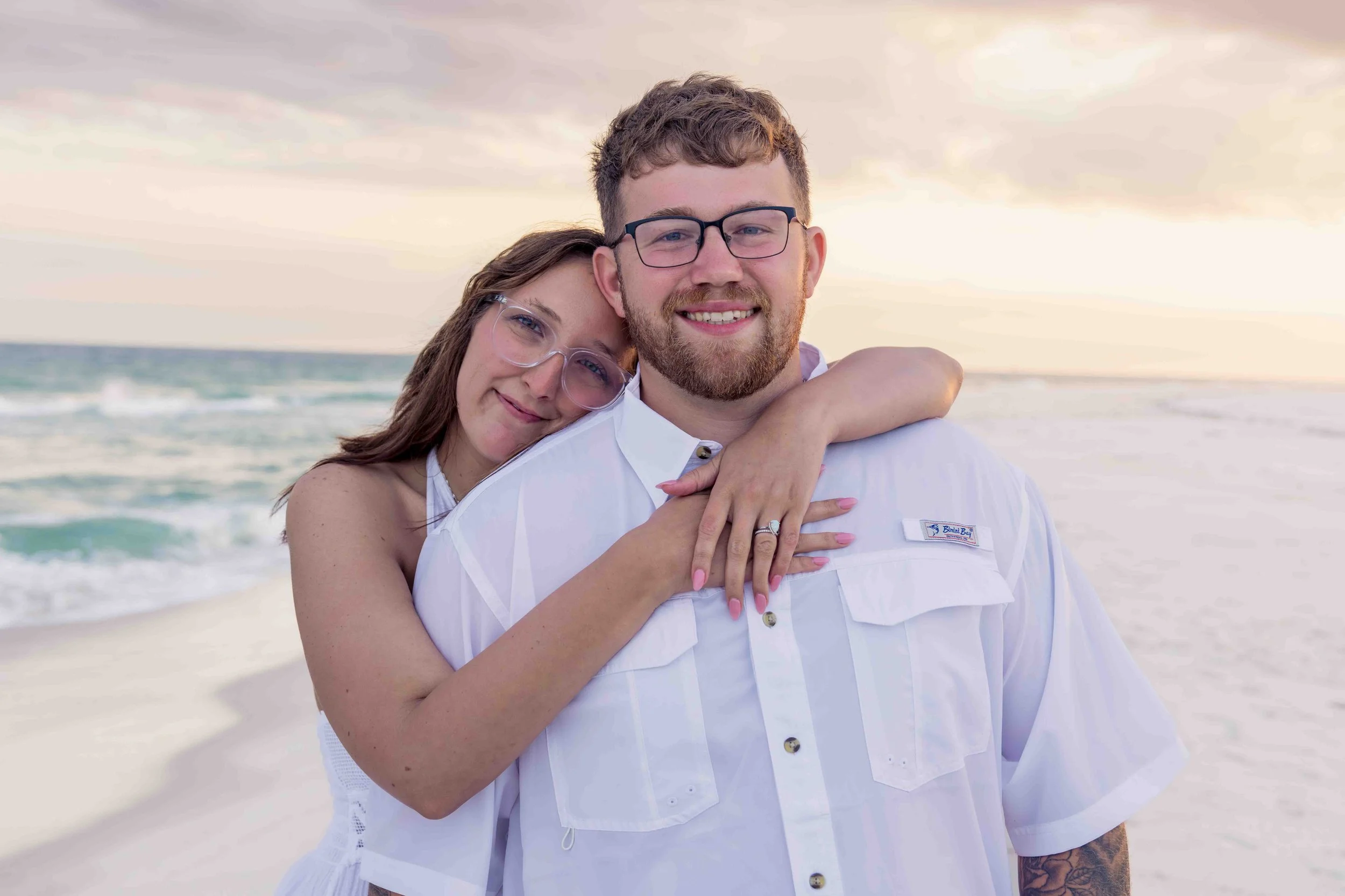 A couple at the beach, with the woman hugging the man from behind, both smiling. The woman is wearing glasses and has her eyes closed, and the man is wearing glasses and a white shirt. The background shows ocean waves and a cloudy sky during sunset o