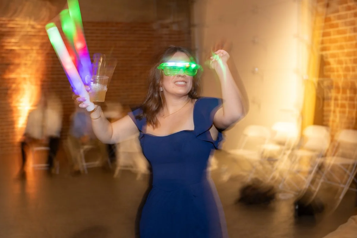 Woman at party wearing green glow glasses, holding glow sticks and a drink, smiling with motion blur in background.