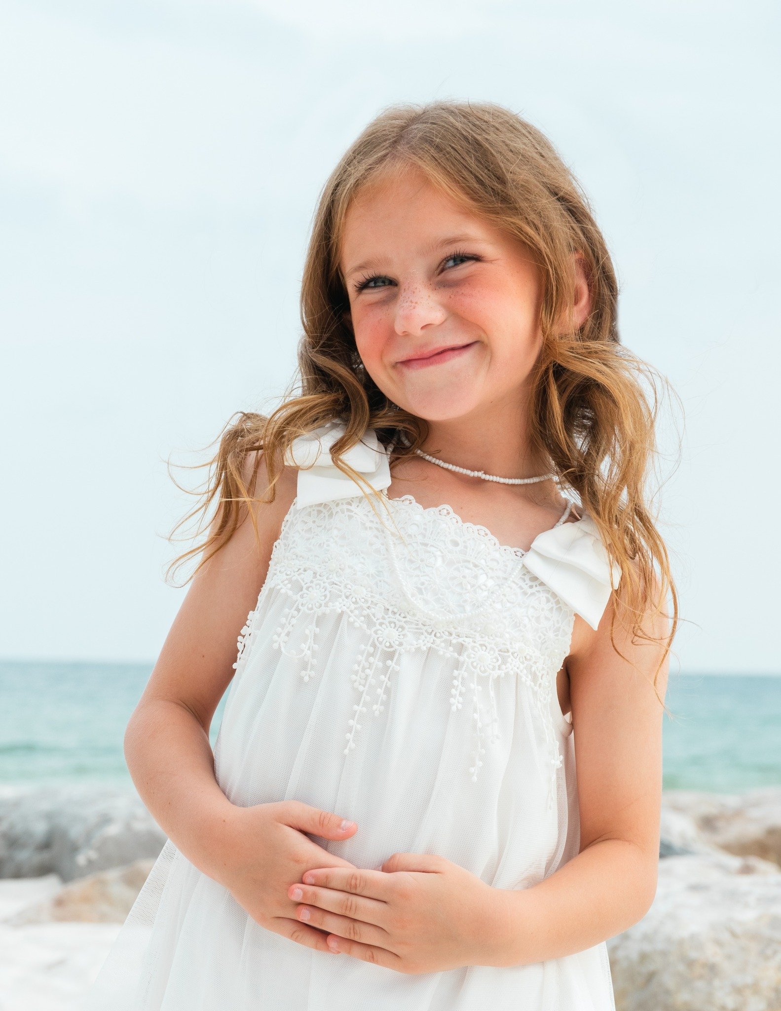 Young girl with curly red hair smiling on the beach, wearing a white dress and necklace, with hands folded over her stomach, ocean in background.