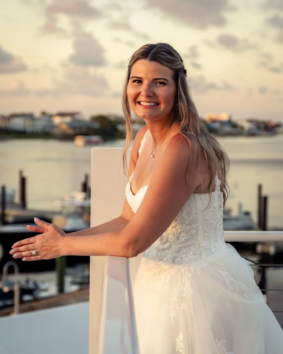 A young woman in a white wedding dress smiling, standing outdoors near water with boats and buildings in the background during sunset.