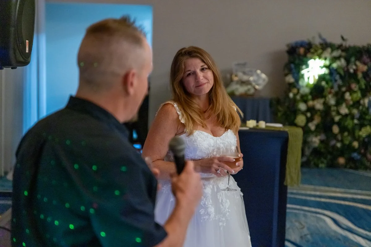 A woman in a white dress holding a wine glass looks at a man giving a speech at a celebration or wedding reception.