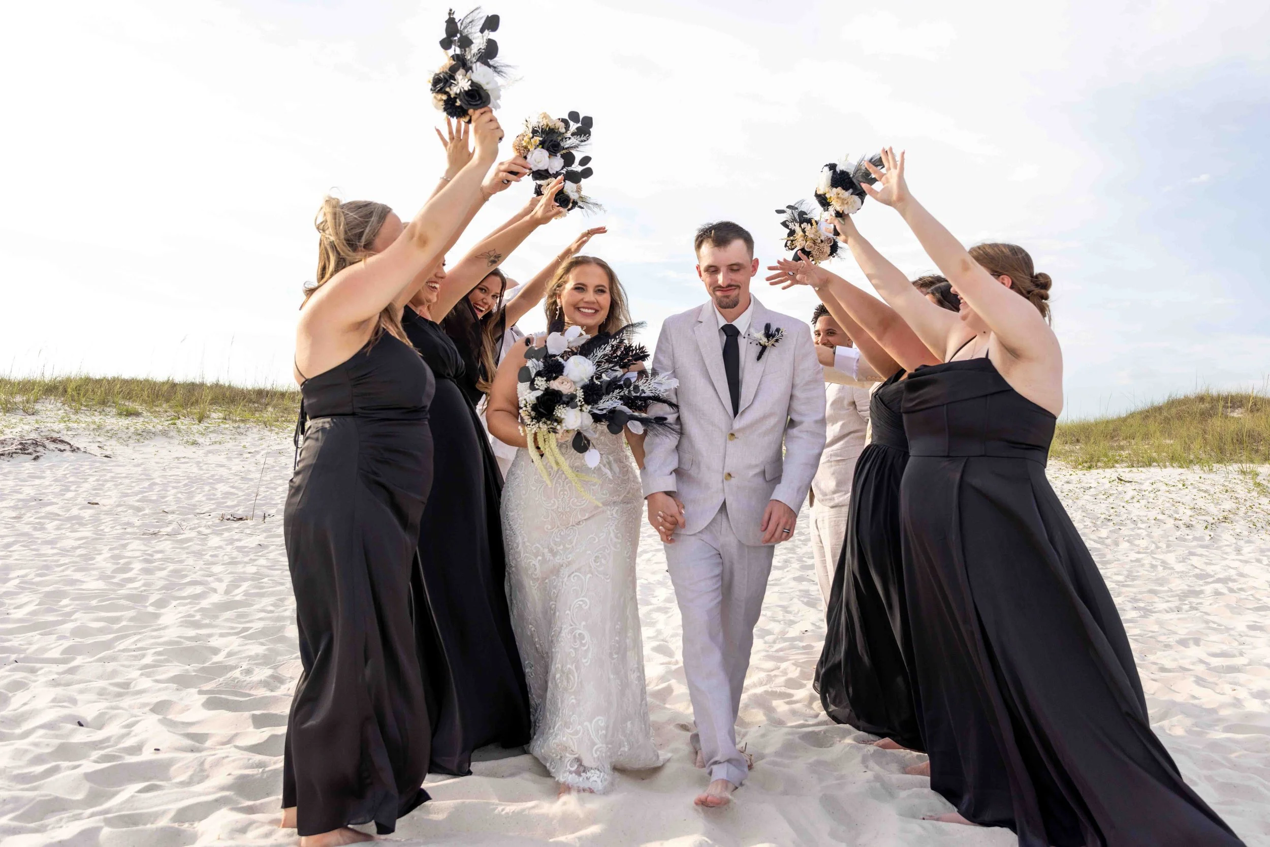 A bride and groom walking hand in hand on a sandy beach, surrounded by bridesmaids in black dresses holding bouquets overhead, celebrating their wedding.