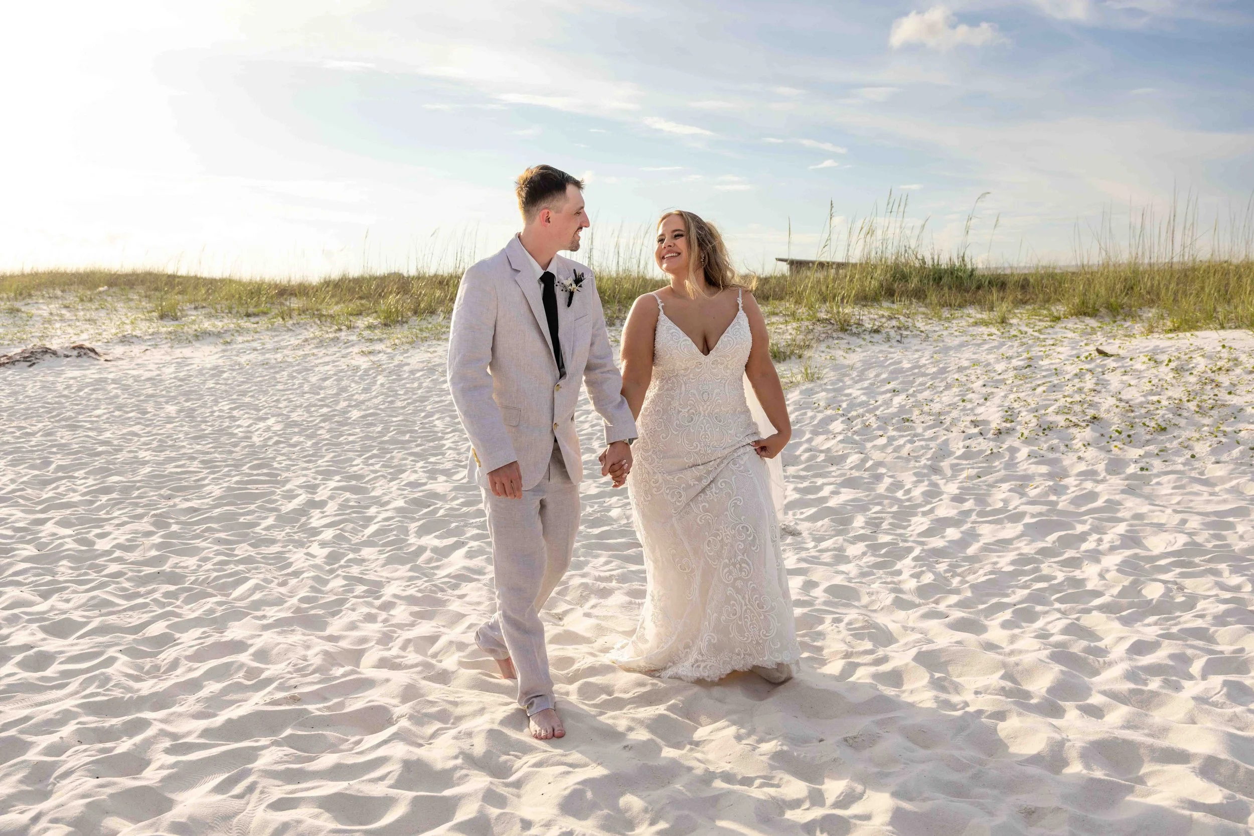 A bride and groom holding hands walking on a sandy beach, smiling at each other, with grassy dunes and partly cloudy sky in the background.