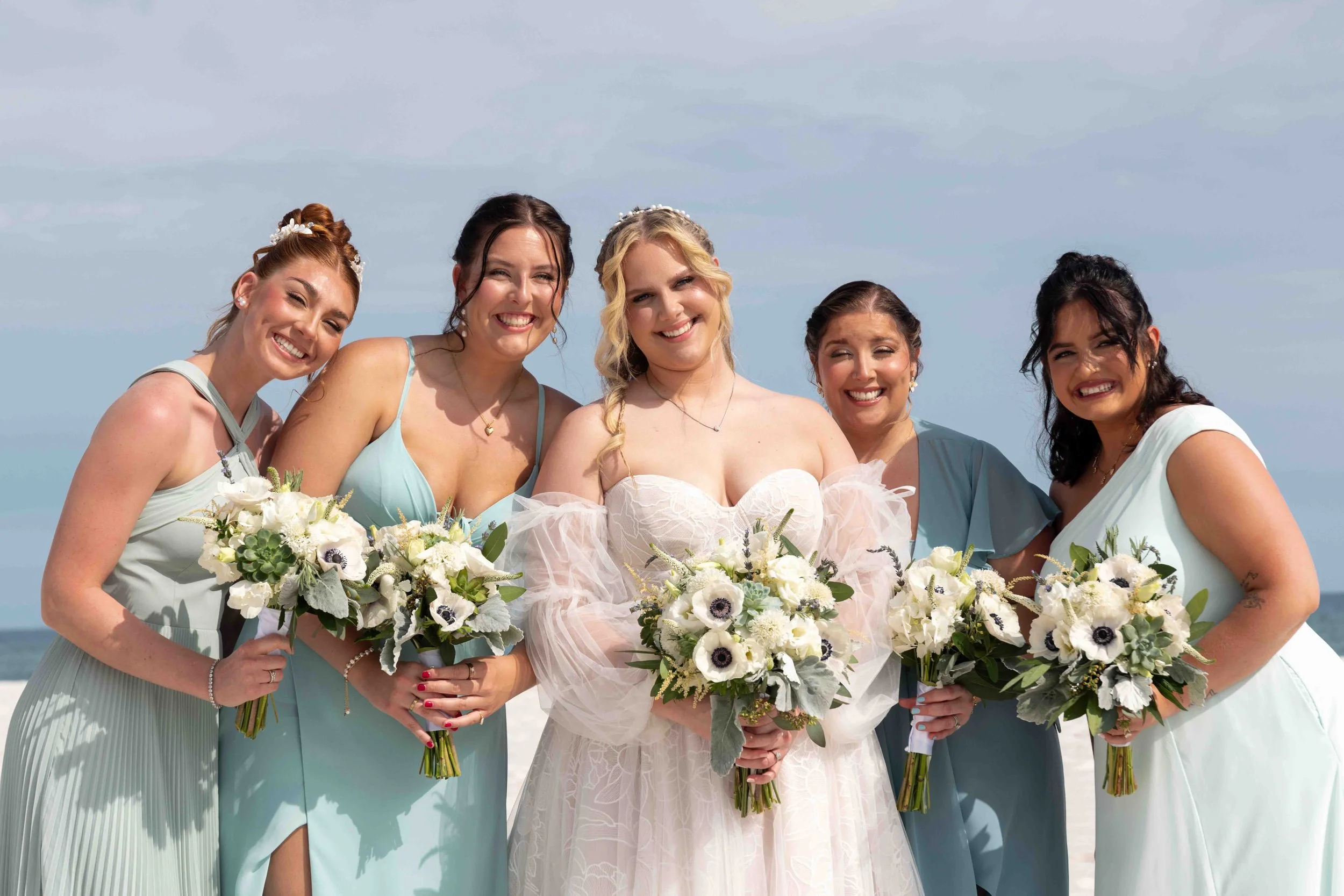 Group of five women at a beach, dressed in pastel-colored dresses, holding bouquets of white flowers, smiling for a photo during a wedding celebration.