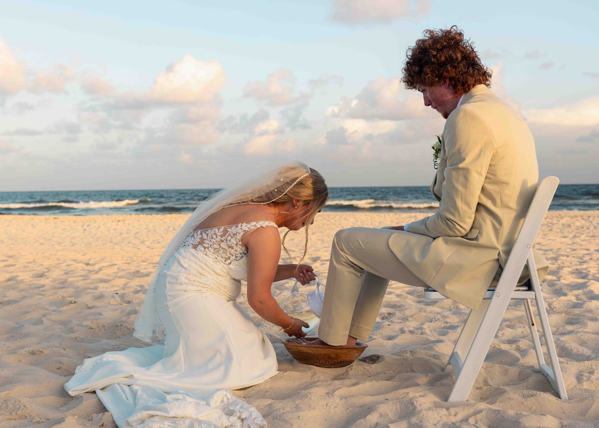 A woman in a wedding dress kneels on the sand in front of a seated man during a wedding ceremony on the beach, with the ocean and sky in the background.
