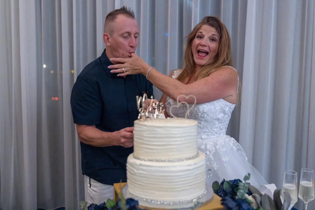 A bride and groom at their wedding reception, with the bride playfully touching the groom's face near a wedding cake.