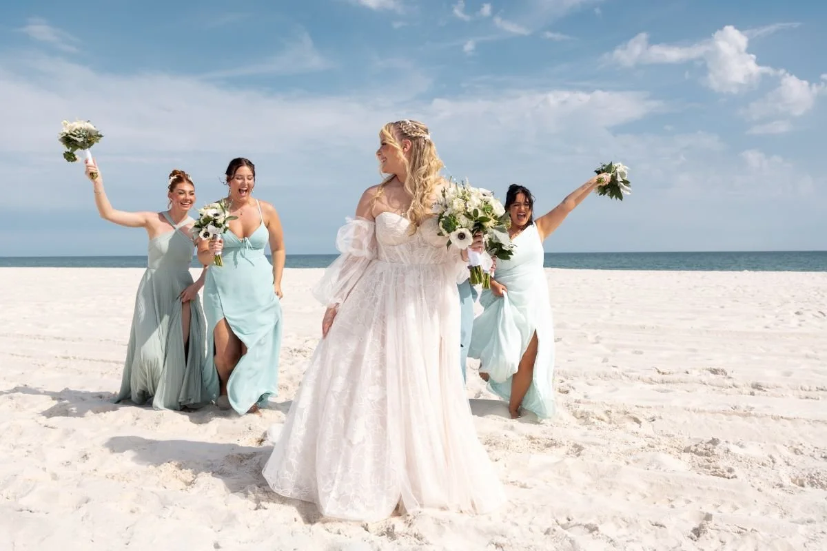 A bride in a white lace wedding dress holding a bouquet of flowers stands on a sandy beach, looking at four bridesmaids who are celebrating with bouquets in their hands.