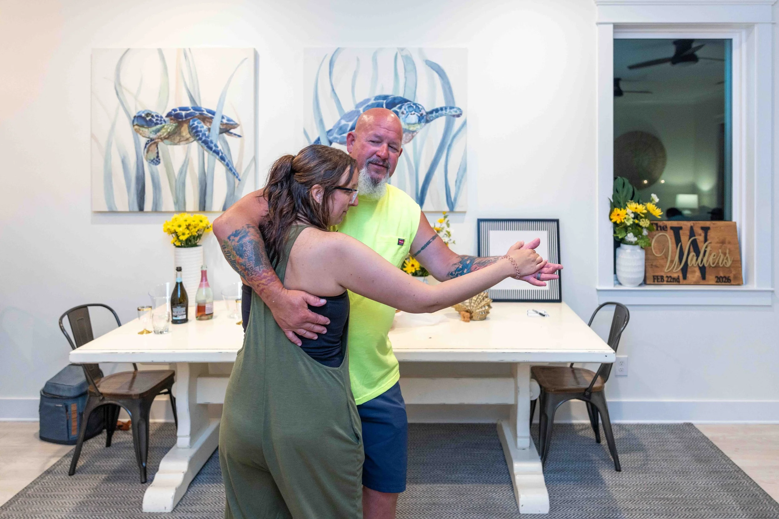 Two people hug in a dining room, one with tattoos and a green sleeveless top, the other with a shaved head and beard, wearing a bright yellow sleeveless shirt. Artworks of sea turtles are on the wall, and there are flowers and signs with the text 'Th