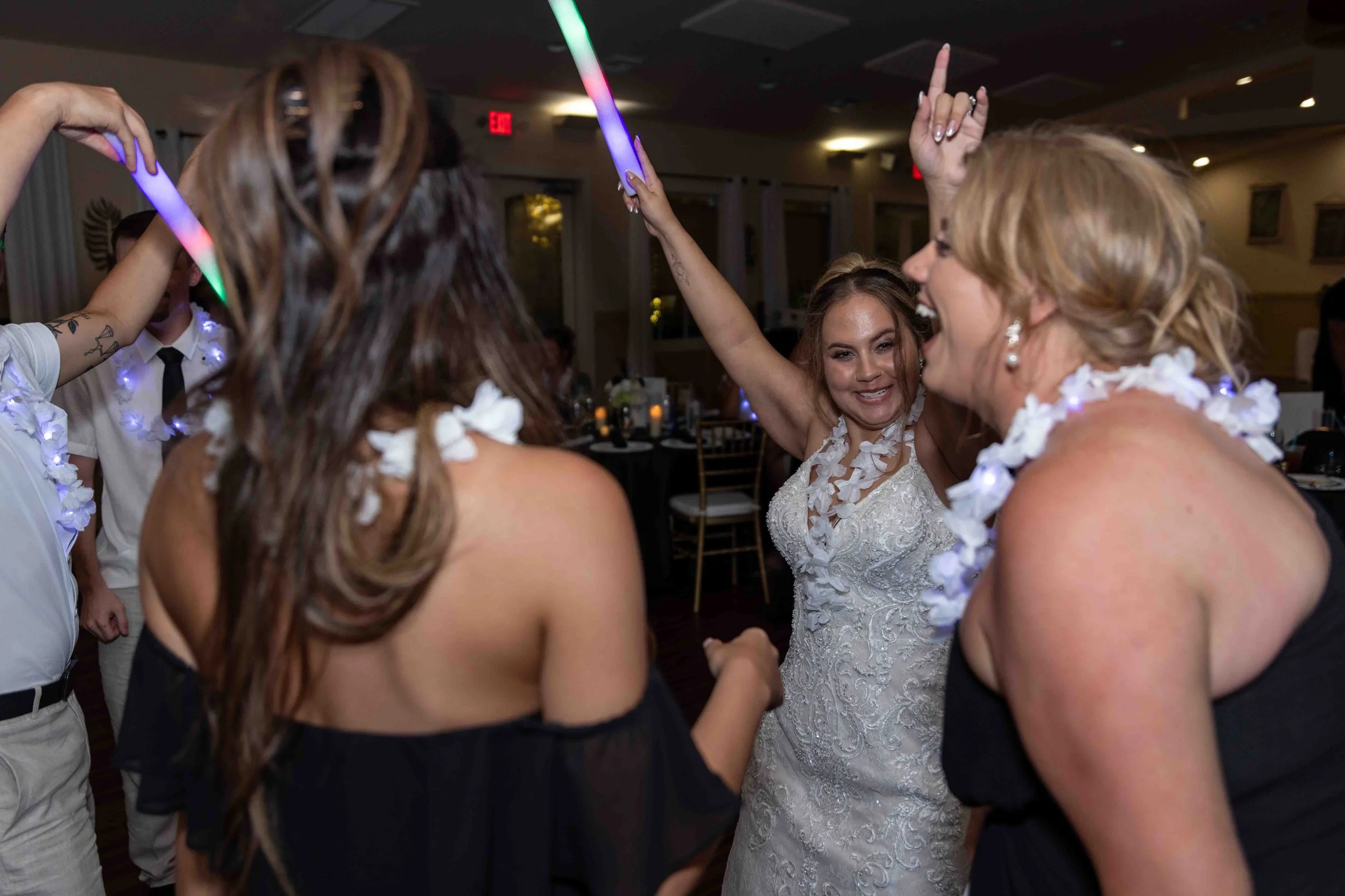 People dancing at a wedding reception, including a bride in a white lace dress and women wearing leis, in a dimly lit banquet hall.