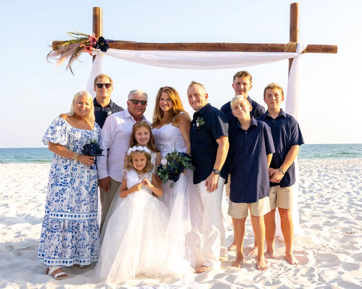 A group of ten people standing on a sandy beach under a wooden arch decorated with fabric and flowers, celebrating a wedding.