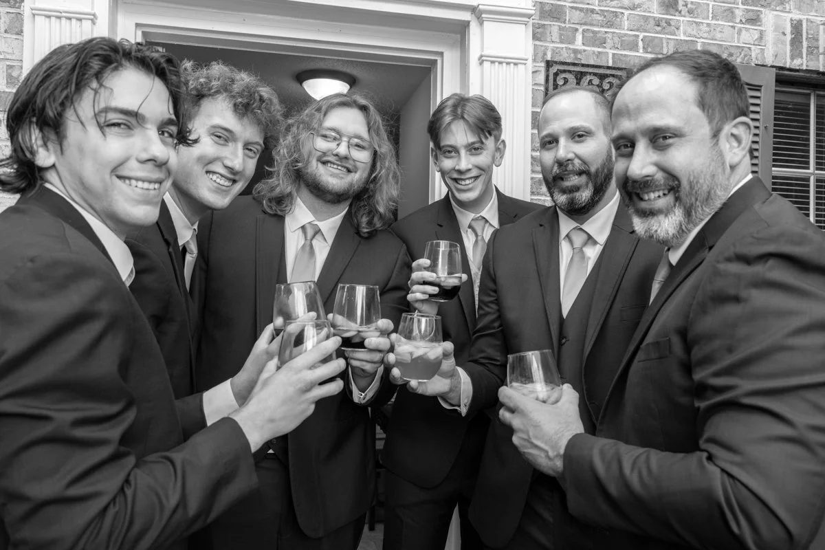 Group of six men in suits smiling and holding glasses of wine at a social gathering.