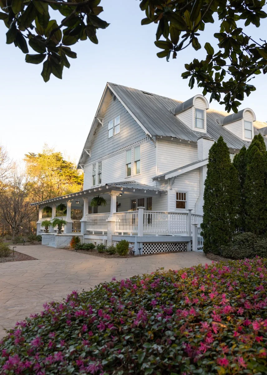 A white, multi-story house with a metal roof, surrounded by trees and bushes, with a porch decorated with hanging plants and potted flowers.