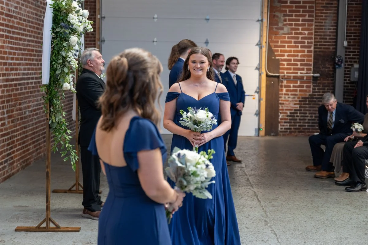 A wedding ceremony with bridesmaids dressed in navy blue gowns, one holding a bouquet of white flowers, standing in front of a brick wall and a floral arch. People seated on benches are visible in the background.