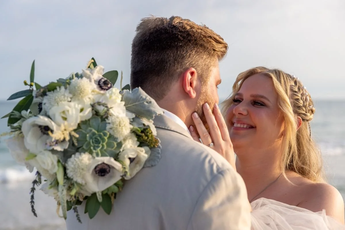 A couple dressed in wedding attire, holding each other on a beach with waves in the background, with the woman smiling and touching the man's face.