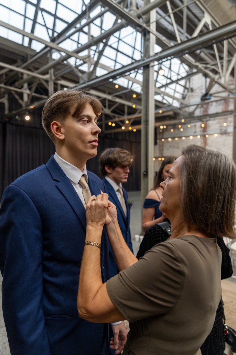 A young man in a blue suit and tie standing up as an older woman, possibly officiating a wedding, adjusts his collar or lapel. The setting is an industrial-style venue with metal beams and string lights, with other people dressed formally visible in 