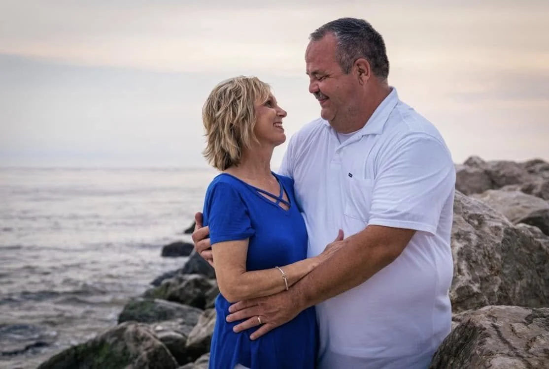 A couple hugging and smiling on a rocky beach at sunset.