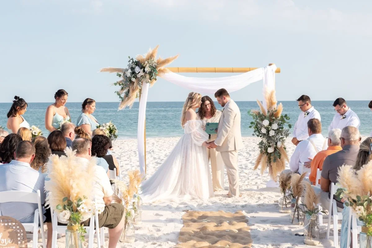 Beach wedding ceremony with a bride and groom exchanging vows underneath an arch decorated with flowers and pampas grass, with guests seated on both sides, and the ocean in the background.