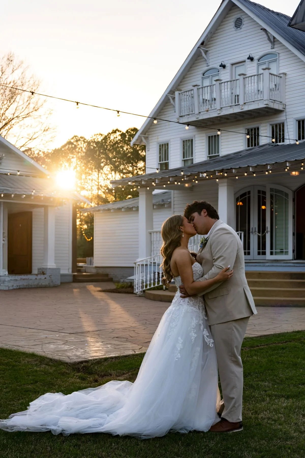 Bride and groom kissing outdoors at sunset, in front of a white house with string lights and a balcony, wearing wedding attire.