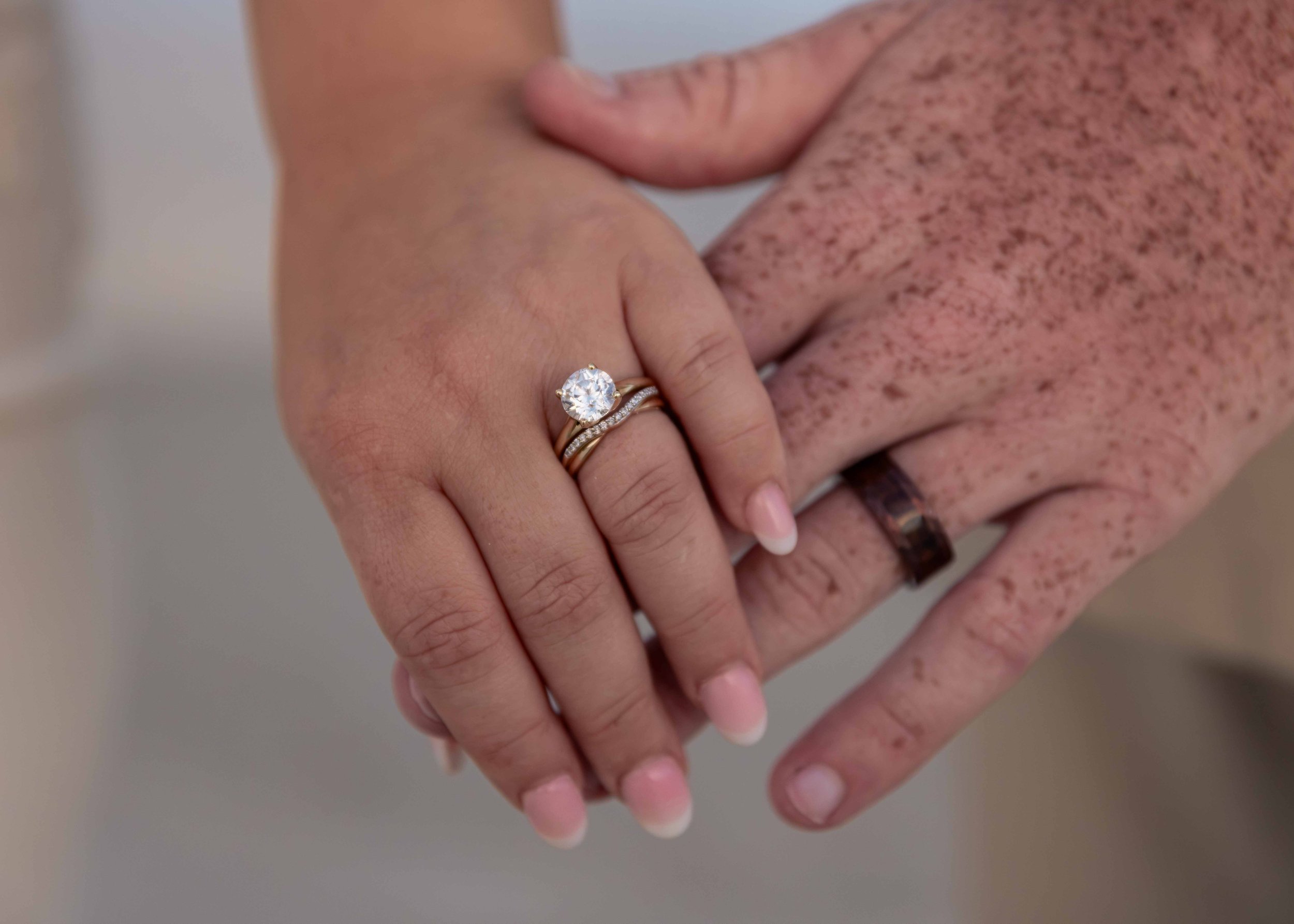 Close-up of two hands holding each other, one with an engagement ring featuring a large round diamond and a band with smaller diamonds, both hands showing some freckles and light skin tones.