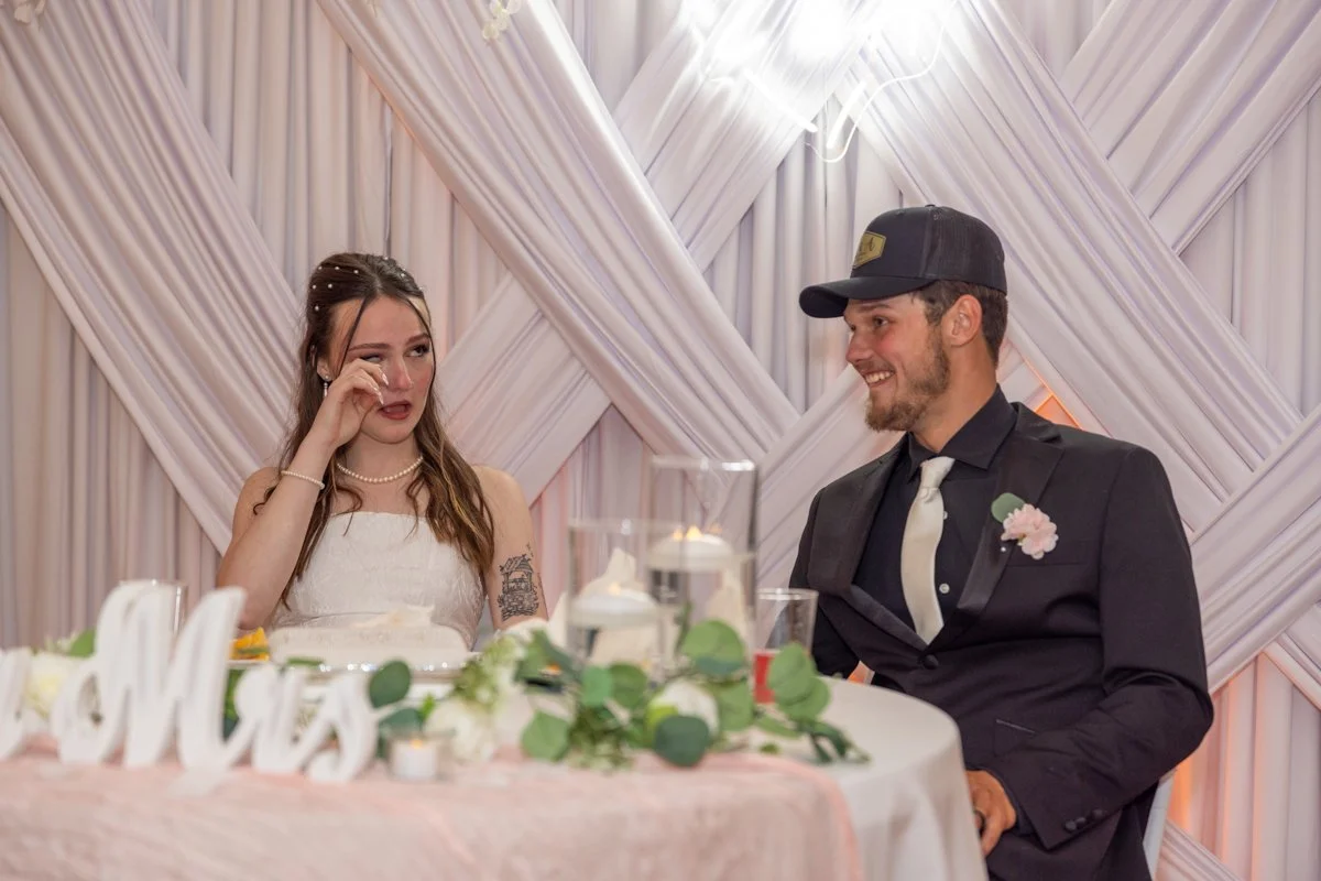 A bride and groom at a wedding reception, seated at a decorated table with a candle centerpiece, greenery, and a "Mr and Mrs" sign, with a backdrop of draped fabric and pink lighting.