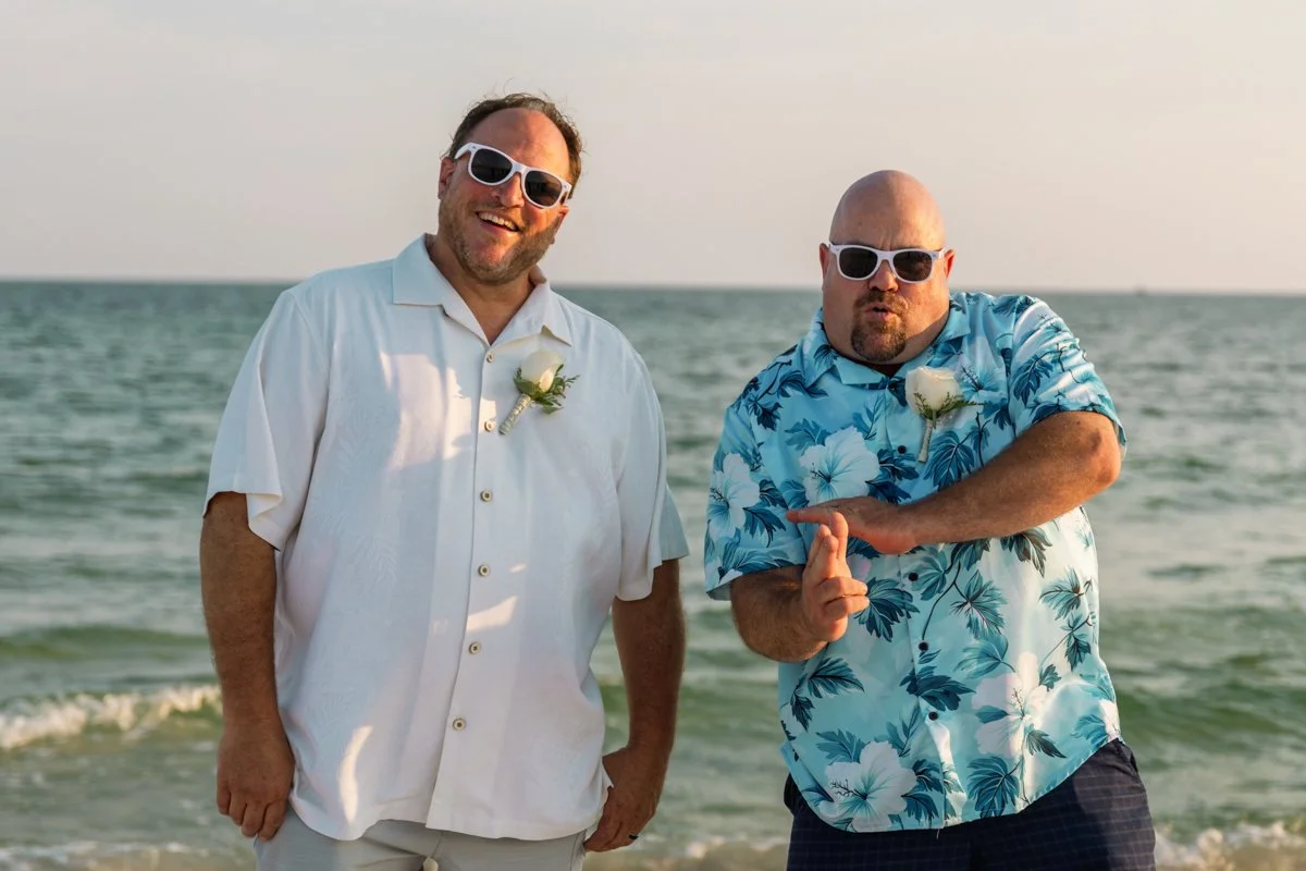 Two men with sunglasses and boutonnières on their shirts standing on a beach with the ocean in the background, one in a white shirt smiling and the other in a blue Hawaiian shirt striking a playful pose.