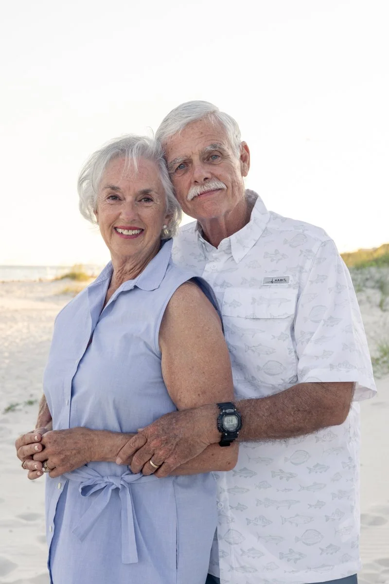 An elderly couple stands on the beach, smiling and embracing each other.