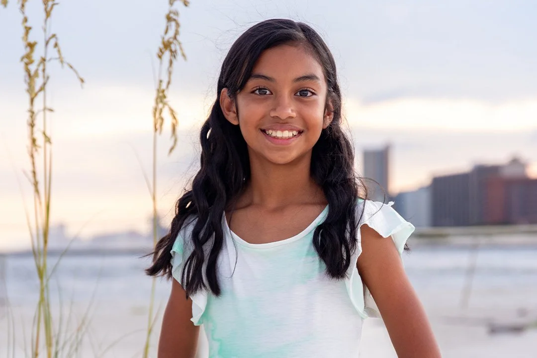 A young girl with long dark hair and a bright smile standing outdoors near water with city buildings in the background at sunset.