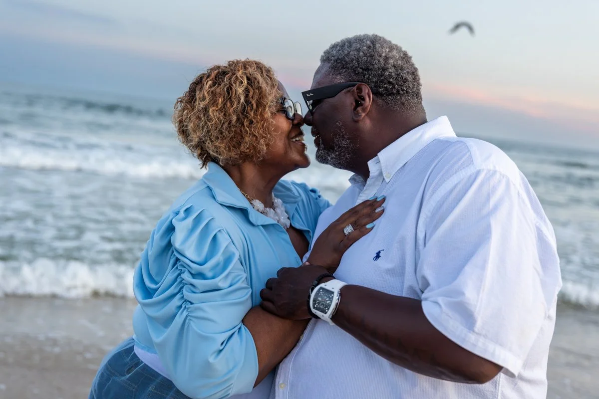 An older couple with gray hair and glasses smiling and touching foreheads on the beach at sunset, with waves and a bird flying in the background.