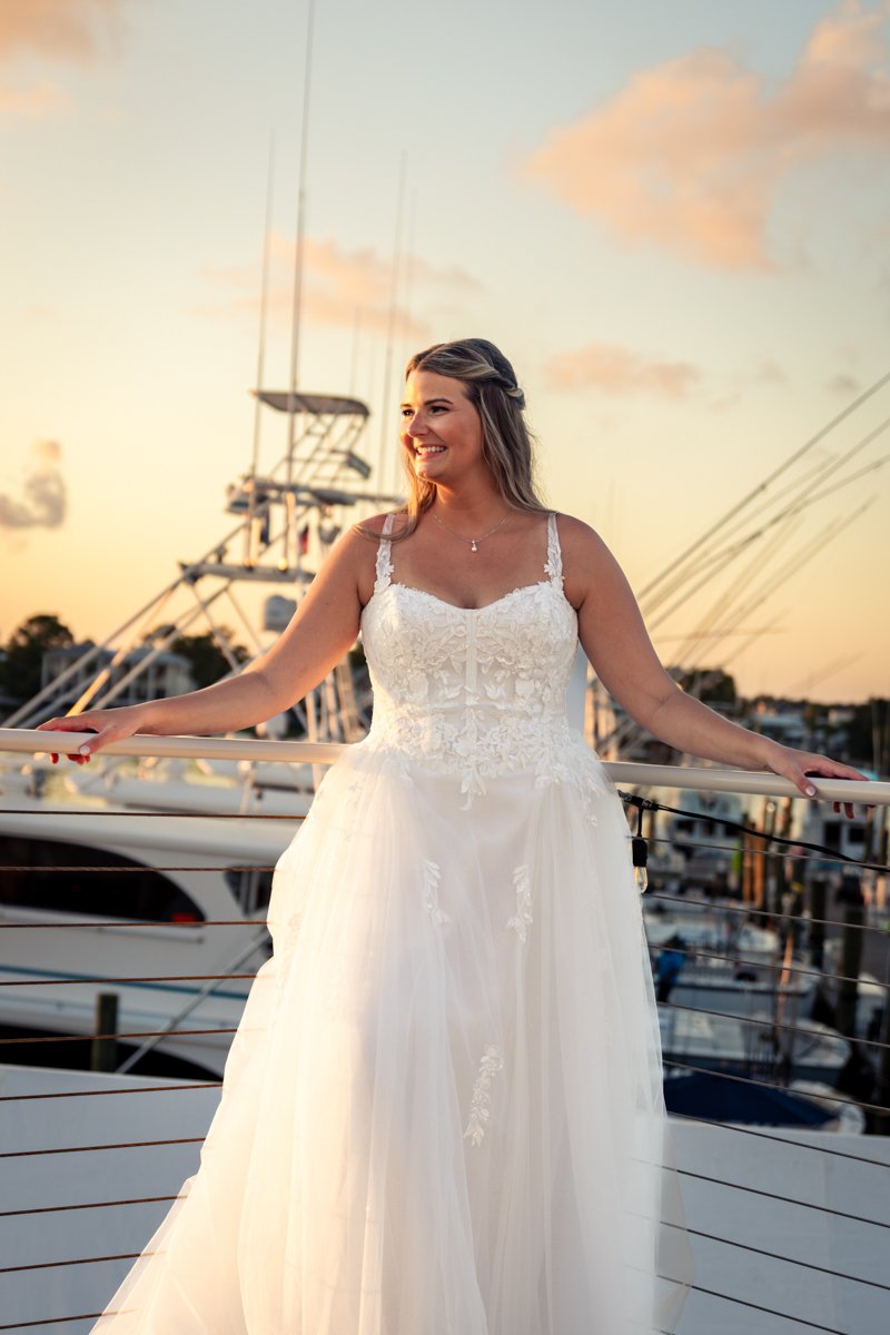 A woman in a white wedding dress standing on a boat with a marina in the background during sunset.