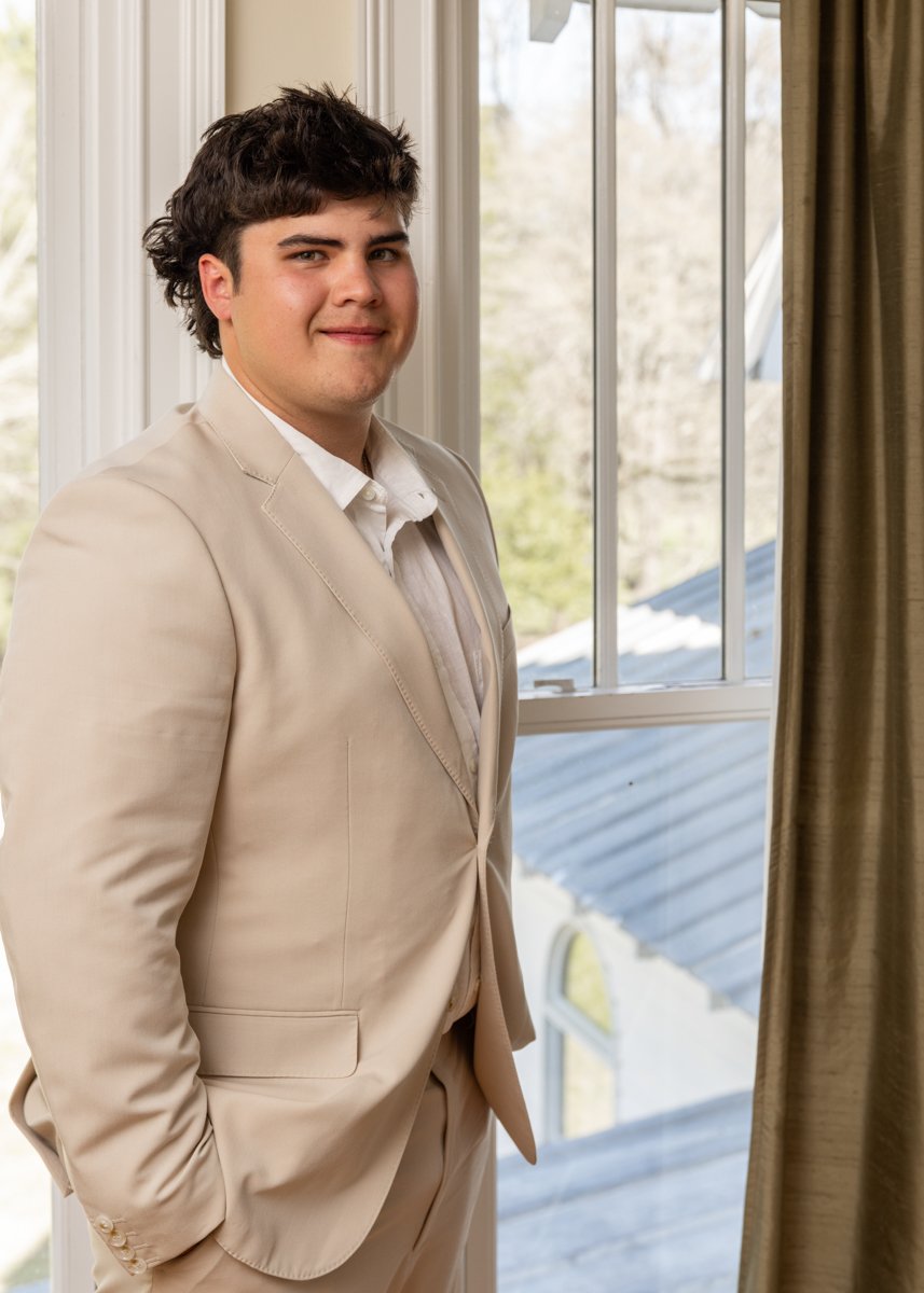 Young man in a beige suit standing by a window with a view of trees and roof outside.