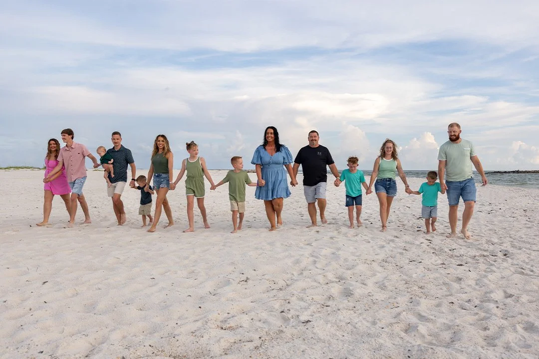 A large family holding hands and walking along a sandy beach under a partly cloudy sky.